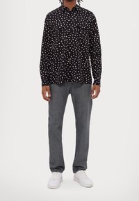 Černá košile s dlouhými rukávy a bílým hvězdným vzorem, se dvěma náprsními kapsami, spárovaná se šedými dž íny a bílými teniskami.