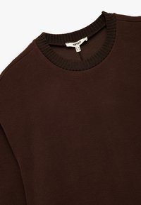Sudadera marrón con cuello redondo y ribete. Textura suave y corte relajado con mangas insertadas, etiqueta visible en el escote.