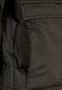 Black puffer jacket material, featuring a smooth texture, thick padding, and a flap pocket on the left side. Stitching is visible along seams.