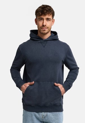 Mann mit kurzen lockigen Haaren, der einen dunkelblauen Hoodie und hellblaue Jeans trägt, Hände in den vorderen Taschen, neutraler Hintergrund.