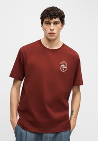 Νεαρός άνδρας με σγουρά μαλλιά, φοράει κόκκινο T-shirt New Balance με το λογότυπο στο στήθος, στέκεται με τα χέρια στις τσέπες μπλε παντελονιού μπροστά σε λευκό φόντο.