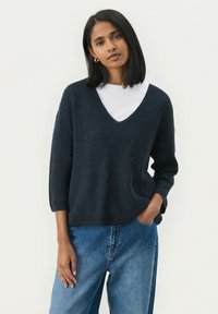 Un pull tricoté bleu marine foncé avec un col en V, des manches 3/4 et une coupe décontractée, porté par-dessus une chemise à col rond blanche. Un jean bleu taille haute.