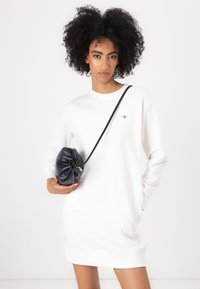 Abito bianco a maniche lunghe con colletto tondo e un piccolo logo. Borsa rotonda in pelle nera e blu con chiusura a cordoncino.