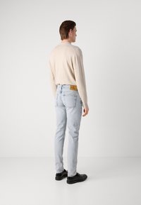 Világos bézs kötött pulóver, slim-fit világoskék farmernadrág hátulsó zsebekkel, fekete cipők, a modell hátulról látható egy sima háttér előtt.