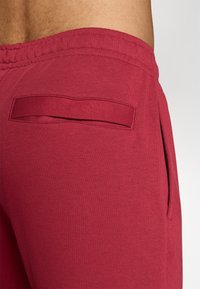 Sarkani kokvilnas sporta bikses ar elastīgu jostu, sānu kabatu un gludu auduma tekstūru. Aizmugurējā dizainā ir horizontāla kabata.