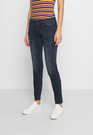 Dark blue skinny jeans made of denim, featuring five pockets, subtle fading, and a fitted design, worn with white sneakers.