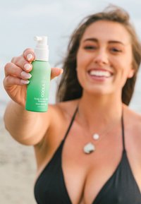 Zielona plastikowa butelka sprayu z etykietą "COOLA Scalp & Hair Mist Sunscreen". Posiada białą nasadkę pompkową i gładką powierzchnię. Pojemność 2 fl oz/60 ml.