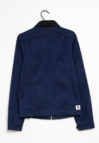 Mörkblå jacka med mörk krage, med knappmanschetter och en logotyp-patch vid fållen. Tillverkad av texturerad tyg, hängande på en krok.