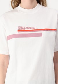 Bijela pamučna majica s okruglim izrezom i kratkim rukavima. Na prednjoj strani prikazan je logo "KARL LAGERFELD JEANS" u ružičastim i crvenim prugama.