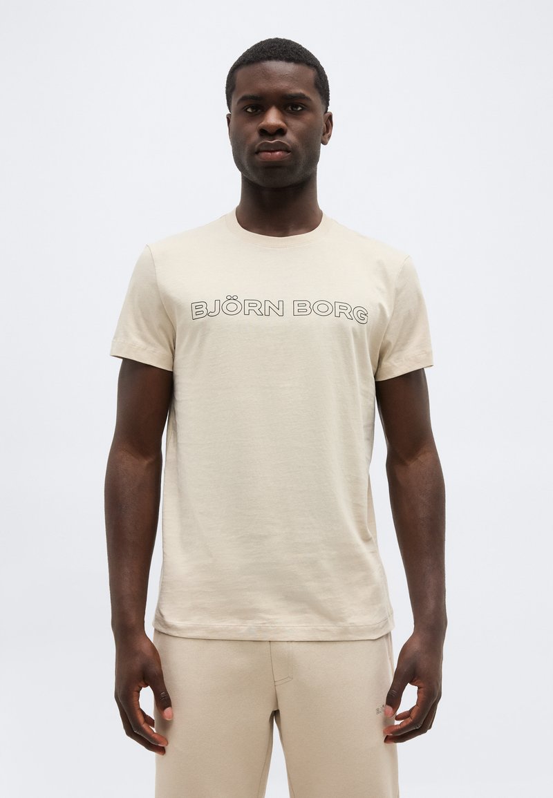 Νεαρός άνδρας που φοράει μπεζ μπλουζάκι Björn Borg και ταιριαστό παντελόνι, στέκεται μπροστά σε απλό φωτεινό φόντο, κοιτώντας προς τα εμπρός.