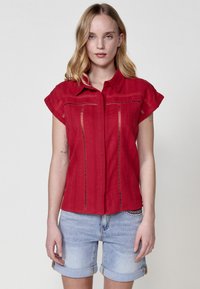 Rode korte mouwen shirt met een knoopsluiting aan de voorkant, voorzien van gedetailleerde stiksels en uitgesneden accenten. Gecombineerd met lichte spijkerbroek shorts.