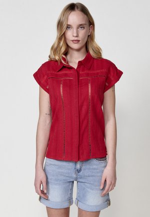 Red short-sleeve shirt with a front button closure, featuring detailed stitching and cut-out accents. Paired with light denim shorts.