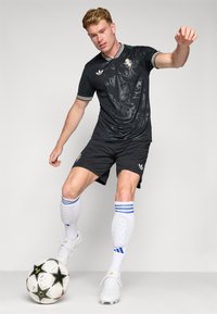 Sort atletisk trøje med grafisk design, parret med sorte shorts. Hvide sokker med blå striber. Spiller i bevægelse med en fodbold.