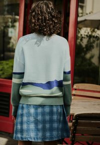 Eine Person mit lockigem Haar, die einen hellblauen und grünen Pullover sowie einen blauen Karottrock trägt, steht vor einer rotgerahmten Glastür in der Nähe von Holztischen und -stühlen.