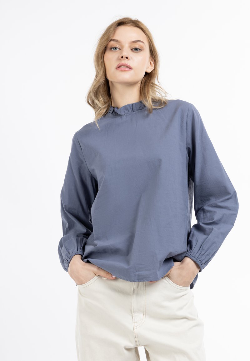Lichtblauwe blouse met een hoge ruches kraag, lange mouwen en elastische manchetten; gemaakt van gladde stof; gestyled met crèmekleurige broek.