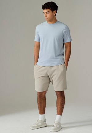 Joven de pie con las manos en los bolsillos, vistiendo una camiseta azul claro, pantalones cortos beige, calcetines blancos y zapatillas blancas, mirando hacia un lado.