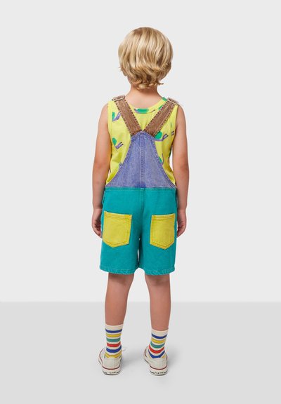 Colorful denim overalls with adjustable brown straps, blue top, green shorts, and large yellow pockets; paired with striped socks and white shoes.