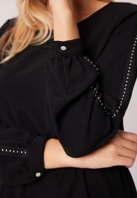 Czarna bluzka z bufkami, z detalami w postaci srebrnych ćwieków na rękawie oraz okrągłym mankiecie z srebrnym guzikiem. Gładka faktura materiału.