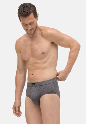 Hombre sin camiseta ajustando unos calzoncillos grises con patrón, de pie contra un fondo blanco liso, mirando hacia abajo con expresión neutral.