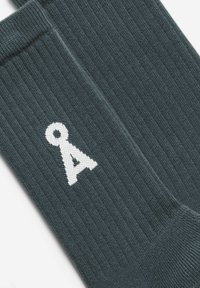 Dunkelgraue, gerippte Socken mit einem weißen Logo. Der Stoff hat eine glatte Textur mit leichtem Glanz, und das Design ist minimalistisch.