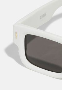 Gafas de sol blancas con montura gruesa, lentes oscuros y un pequeño acento dorado en el lateral. El diseño es angular y moderno.