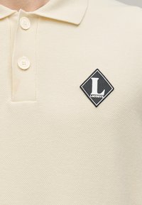 Polo beige à manches courtes en tissu texturé, avec un patch logo noir en forme de losange orné d'un « L » blanc sur le côté gauche de la poitrine. Fermeture par trois boutons.