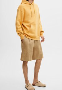 Sudadera amarilla con capucha y cordón ajustable, bolsillo delantero, combinada con pantalones cortos marrón claro y zapatos de lona beige.