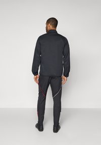 Sort træningsjakke med elastiske manchetter, parret med sorte bukser med røde og hvide diagonale striber. Struktureret stof, funktionelt design.