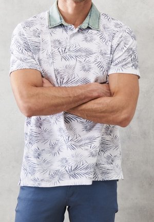 Uomo che indossa una polo bianca a maniche corte con motivi di foglie grigie e colletto verde, braccia incrociate, pantaloni blu, sfondo grigio chiaro.