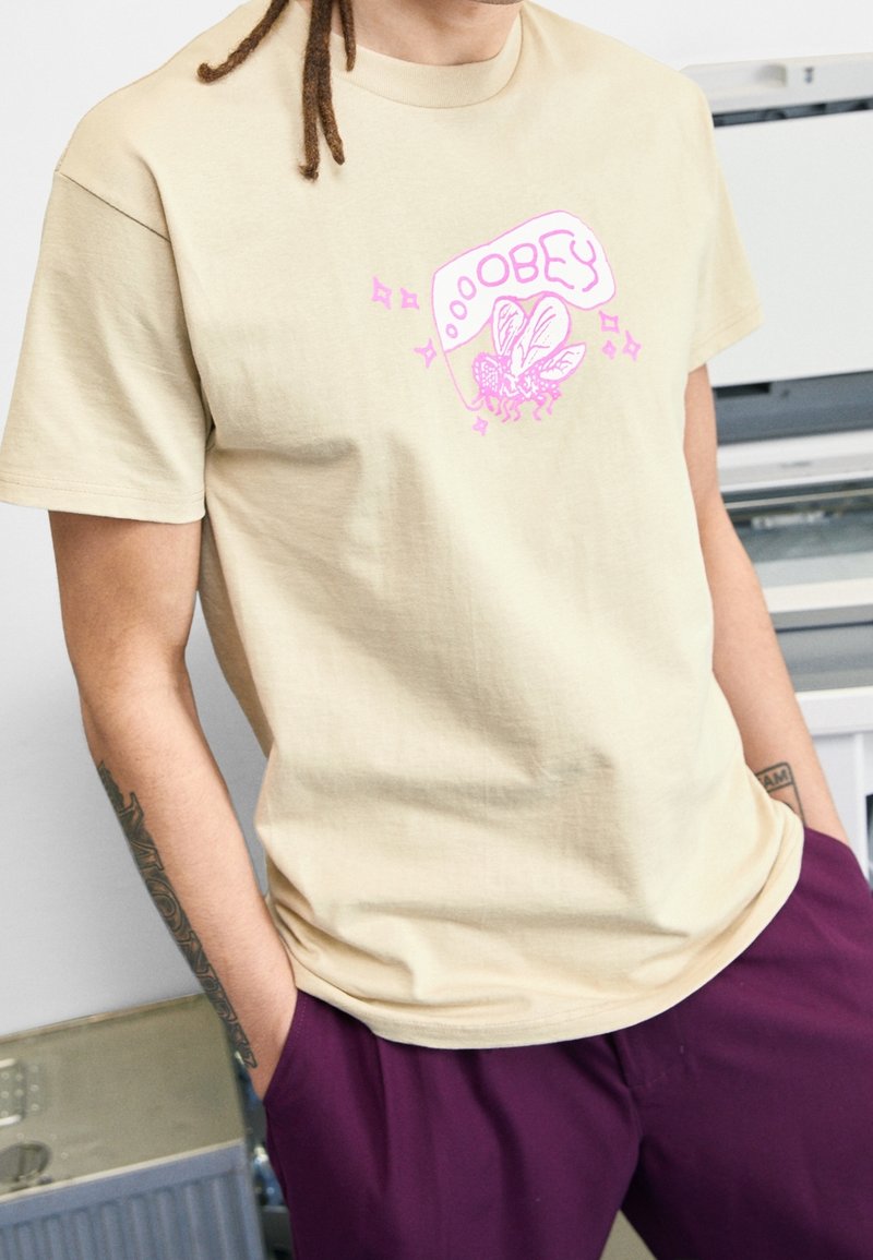 Beige t-shirt i bomull med en rosa grafik av en fluga och texten "OBEY." Korta ärmar, rundad hals, avslappnad passform.