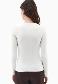 Donna con lunghi capelli castani che indossa orecchini a cerchio d'argento, una maglietta bianca a maniche lunghe aderente e pantaloni marroni scuri, vista da dietro.