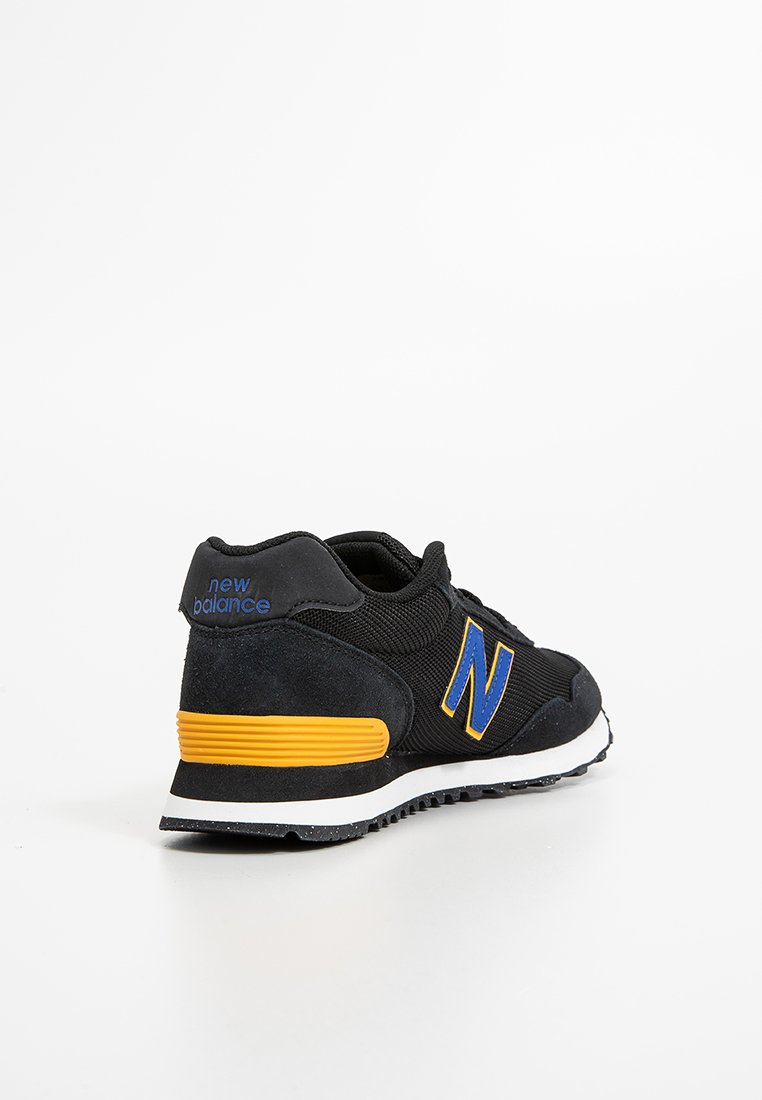 Zapatilla negra con parte superior de textil y ante, con una cinta de acento amarilla y un logo azul. Diseño clásico, suela de goma con agarre texturizado.