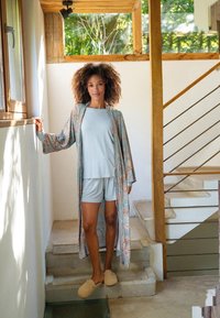 Blumenmuster-Kimono in Blau und Koralle über einem hellblauen Top und Shorts. Beigefarbene Hausschuhe auf Betonstufen mitHolzdetails.