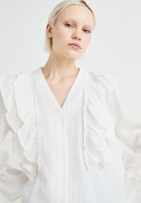 Hvit bluse med volang foran, V-hals og knappelukking. Laget av lett, teksturert stoff med lange ermer og elastiske mansjetter.