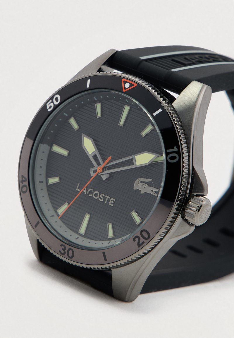 Lacoste horloge met zwarte wijzerplaat, lichtgevende groene uurmarkeringen en wijzers, rode secondewijzer, zwarte draaibare bezel en zwart rubberen band.