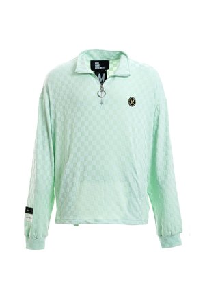 Joven de pie, vistiendo un jersey de manga larga a cuadros verde menta claro con cremallera, pantalones cortos a juego con ribete negro, calcetines blancos y zapatillas beige.