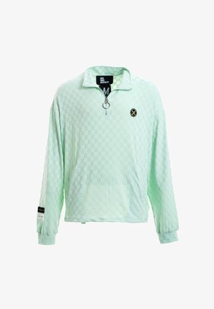 Joven de pie, vistiendo un jersey de manga larga a cuadros verde menta claro con cremallera, pantalones cortos a juego con ribete negro, calcetines blancos y zapatillas beige.
