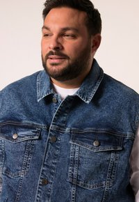 Džínová bunda ve středně modré barvě, s špičatým límcem, dvěma kapsami na prsou, stříbrnými cvoky a viditelnými švy.