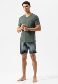 Groene T-shirt met korte mouwen en V-hals, voorzien van een borstzak, gecombineerd met marineblauwe gestreepte shorts. Model staat blootsvoets tegen een effen achtergrond.