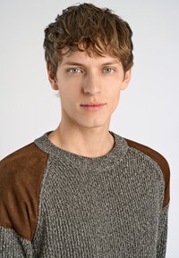 Suéter de punto gris con parches de ante marrón en los hombros; presenta un escote redondo y una tela texturizada. El diseño es casual y ajustado.