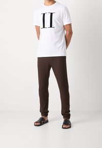 Camiseta blanca de algodón con gráfico negro "II", combinada con pantalones ajustados marrón oscuro y sandalias negras tipo sandalia. Diseño sencillo y casual.