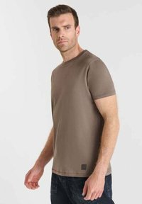 Uomo in camicia corta marrone semplice e pantaloni scuri, in piedi e che guarda leggermente di lato su uno sfondo semplice.