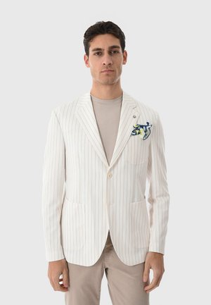 Uomo che indossa una giacca beige a righe sottili con fazzoletto da taschino blu e giallo sopra camicia e pantaloni taupe, in piedi davanti a uno sfondo grigio chiaro.