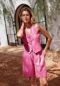 Gilet en lin rose à cinq boutons et shorts assortis. Texture lisse, coupe ajustée et poches latérales. L'arrière-plan présente de la verdure et de l'architecture.