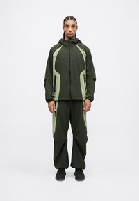 Dvoudílný sportovní outfit v tmavě zelené látce se světle zelenými bočními akcenty. Obsahuje kapuci, kapsy na zip a volné kalhoty.