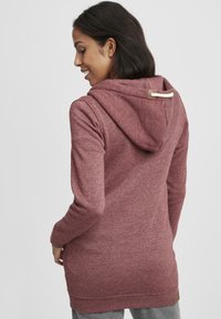 Burgunderfarget hettegenser med teksturert stoff, snorhette og detaljer med sideventilasjon, med en lys beige aksent der hetten festes.