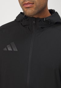 Zwarte sportjack gemaakt van lichtgewicht stof, voorzien van een ritssluiting, verstelbare capuchon en een reliëflogo op de borst.