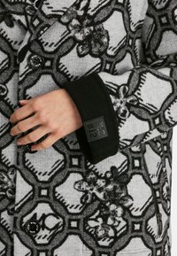 Cappotto a fantasia grigia e nera con motivi floreali, tessuto strutturato, polsini neri e bottoni con marchio, caratterizzato da una zip laterale.