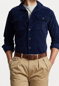 Camisa de pana azul marino con dos bolsillos en el pecho y botones marrones, combinada con pantalones beige con pliegues y un cinturón de cuero marrón.