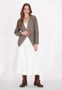 Braune asymmetrische Jacke über einem weißen, bodenlangen Kleid. Das Model trägt braune Stiefel bis zur Mitte der Waden mit Schnallenakzenten. Einfaches Stoffdesign, glatte Textur.